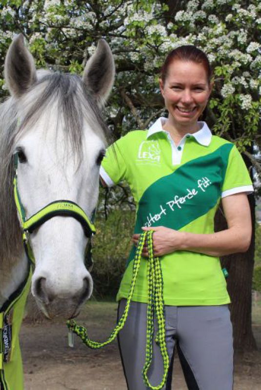 Frau mit Pferd im Freien Eine Frau mit langen roten Haaren steht neben einem grauen Pferd und hält dessen Zügel. Sie trägt ein grünes Polo-Shirt mit einem Logo.