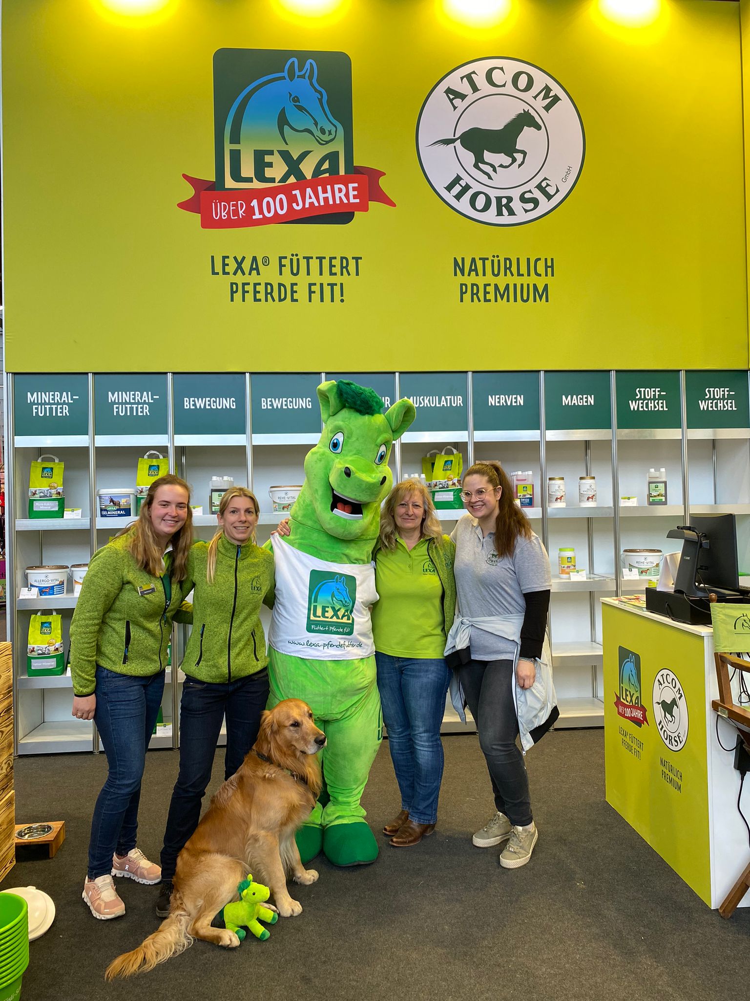 Mitarbeiter von LEXA mit Maskottchen und Hund Gruppe von vier Personen und einem Hund, posierend mit einem Maskottchen in einem Stand für Pferdefutter.